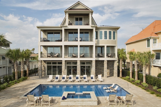 The back exterior of a three-story beach home with several balconies and a large pool with a hot tub attached. There are several lounge chairs on the balconies as well as surrounding the pool area. To the right and left of the house there are palmetto trees.