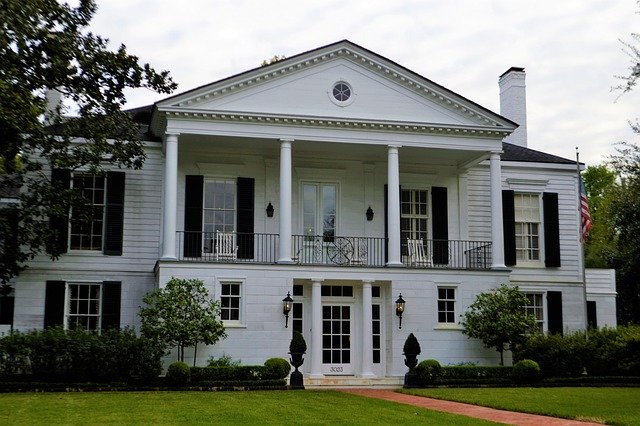 The front exterior of a white, plantation style home with an American flag on the side. There is a brick pathway leading up to the house with green grass to each side.  
