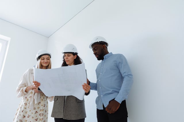 Three adults wearing hard hats are standing shoulder-to-shoulder looking at architectural drawings held by the individual in the center.